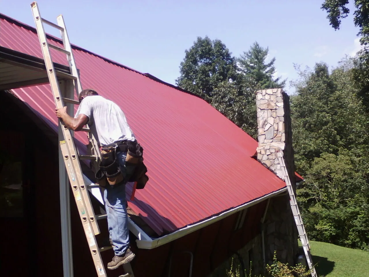 Roofer on a ladder beside a red metal roof and stone chimney — Soffit & Fascia Repair by Roofing Brothers in West Greenwich