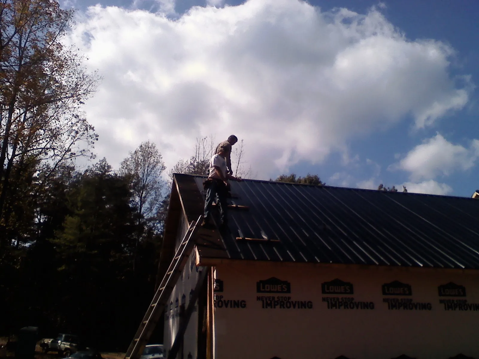 Roofer fastening dark metal panels high against a cloudy sky, from a Roof Inspection job in West Greenwich